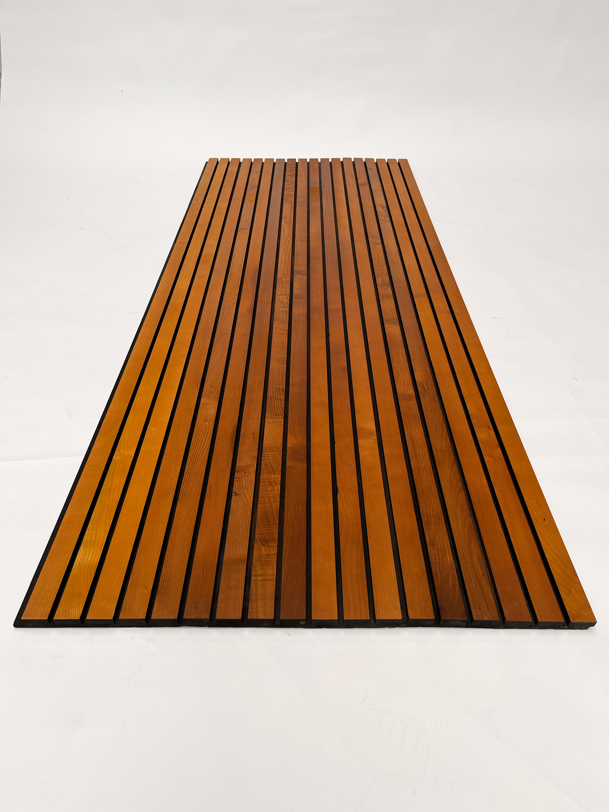 Overhead view of a large wooden facade board with multiple narrow slats, arranged in parallel lines. The board features a warm, natural wood finish with dark grooves between each slat, highlighting its elegant and linear design on a white background.